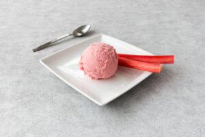 Homemade-Rhubarb-Ice-Cream-A-Truly-Tantalisingly-Tangy-Treat-image-3-two-scoops-of-rhubarb-ice-cream-on-a-white-square-plate-with-teaspoon-with-fresh-rhubarb-stalks-frosted-fusions