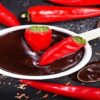 Homemade-Chilli-Chocolate-Ice-Cream-Sweet-and-Spice-Fusion-featured-image-200x600w several red chillis scattered with chunks and shavings of chocolste plus 2 red chillis in white bowl of chocolate sauce frosted fusions