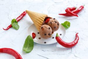 Homemade-Chilli-Chocolate-Ice-Cream-Sweet-and-Spice-Fusion-image-1-2-scoops-of-chocolate-ice-cream-in-cone-on-white-plate-with-fresh-red-chillis-scattered-and-fresh-mint-leaves-frosted-fusions
