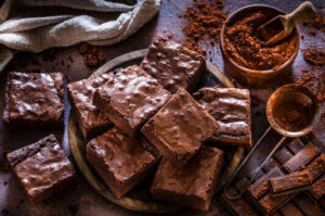 homemade-chocolate-brownie-ice-cream-recipe-image-1-plate-filled-with-chocolate-brownie-squares-with-dish-of-cocoa-sieve-and-chunks-of-chocolate-frosted-fusions