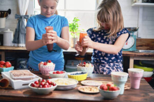 Fun-Easy-And-Healthy-Frozen-Dessert-Recipe-Ideas-For-Kids-image-1-two-girls-in-a-kitchen-with-ingredients-around-them-squeezing-sauce-bottles-frosted-fusions