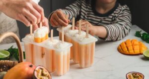 Fun-Easy-And-Healthy-Frozen-Dessert-Recipe-Ideas-For-Kids-image-5-mango-and-yoghurt-lollies-being-pulled-from-mould-with-child-and-adult-hands-fresh-mango-and-fruits-around-frosted-fusions