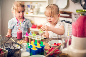 Fun-Easy-And-Healthy-Frozen-Dessert-Recipe-Ideas-For-Kids-image-9-two-younf-girls-taking-fruit-lollies-out-of-moulds-with-various-ingredients-and-cooking-equipment-around-them-frosted-fusions